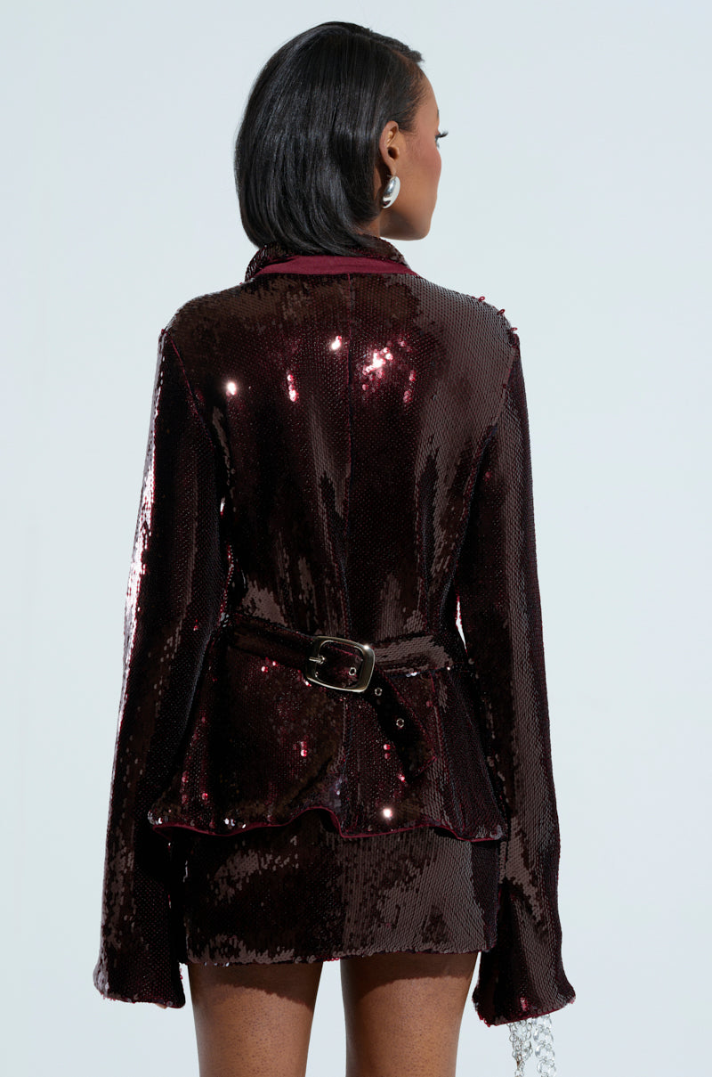 A woman with short dark hair is showcasing the LIGHTS OUT BUTTON DOWN BLOUSE, a shiny dark sequined top paired with a matching skirt that has stretchy mesh panels for comfort. The blouse's belt elegantly cinches her waist as she turns her back to the camera, making her outfit glimmer against the plain light background.