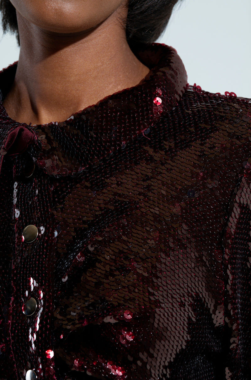 The image showcases a close-up of someone wearing a dark red, sequin-adorned jacket featuring metallic buttons and subtle ruched details. The sequins catch the light beautifully, causing the jacket's surface to shimmer similarly to the LIGHTS OUT BUTTON DOWN BLOUSE, emphasizing the fabric's intricate details.
