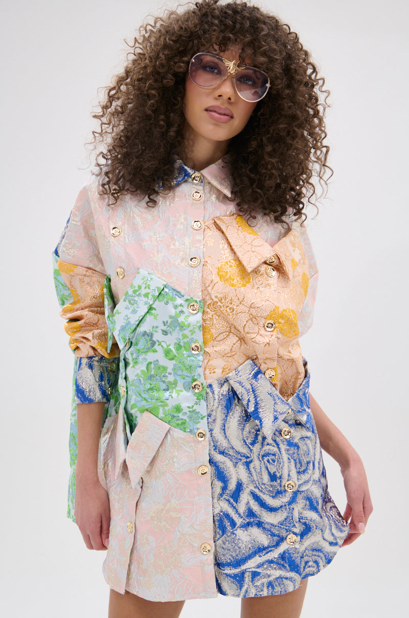 A woman with curly hair wears oversized sunglasses and the MY GIRLFRIENDS SHIRT BROCADE MINI DRESS, a vibrant patchwork mini dress with metallic buttons and fabric bows, standing against a plain light background.