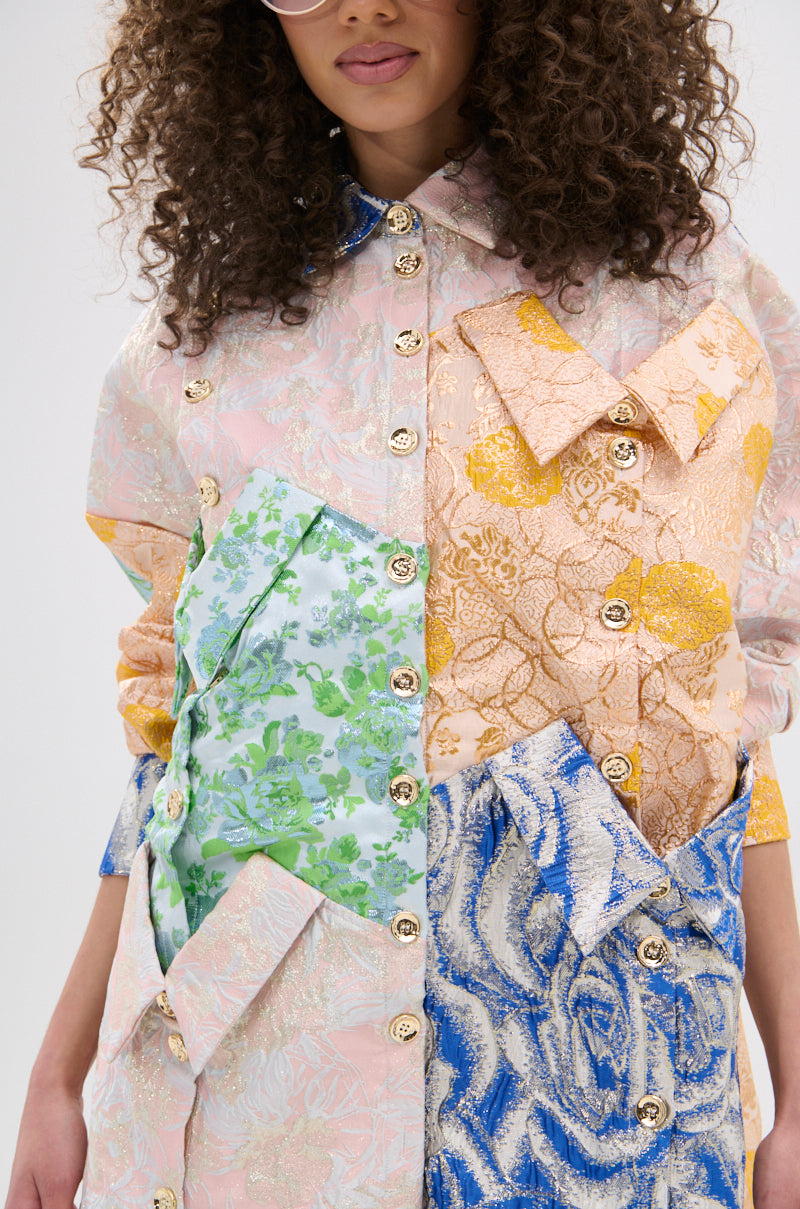 A woman with curly hair wears the MY GIRLFRIENDS SHIRT BROCADE MINI DRESS, featuring a pastel patchwork design, oversized collars, and decorative metallic buttons. Her face is partially visible.
