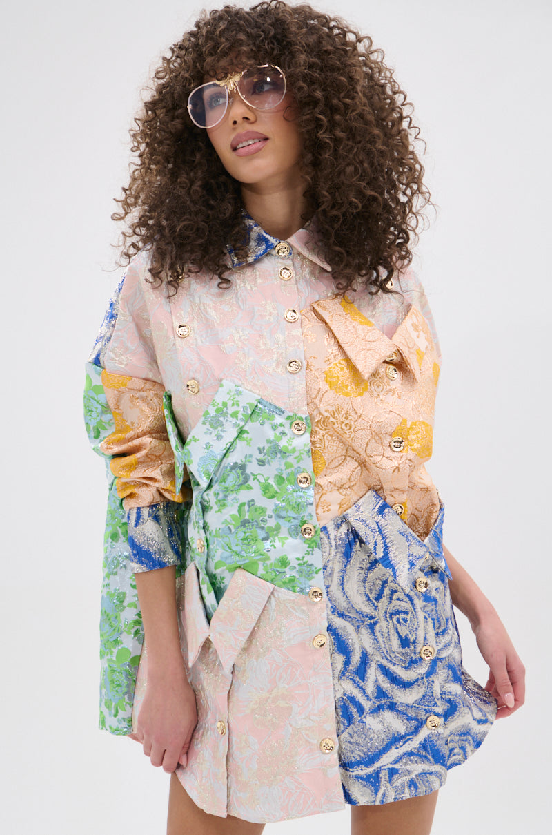 A woman with curly hair and sunglasses wears the MY GIRLFRIENDS SHIRT BROCADE MINI DRESS, a vibrant patchwork mini dress with metallic buttons in pink, green, blue, and gold tones. She poses against a plain white background.