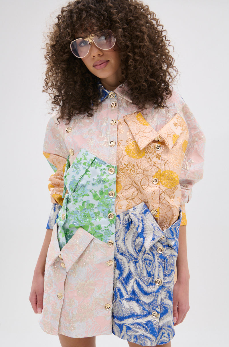 A person with curly hair and oversized sunglasses wears the MY GIRLFRIENDS SHIRT BROCADE MINI DRESS, showing off vibrant patchwork florals and large front pockets, standing against a plain light background.