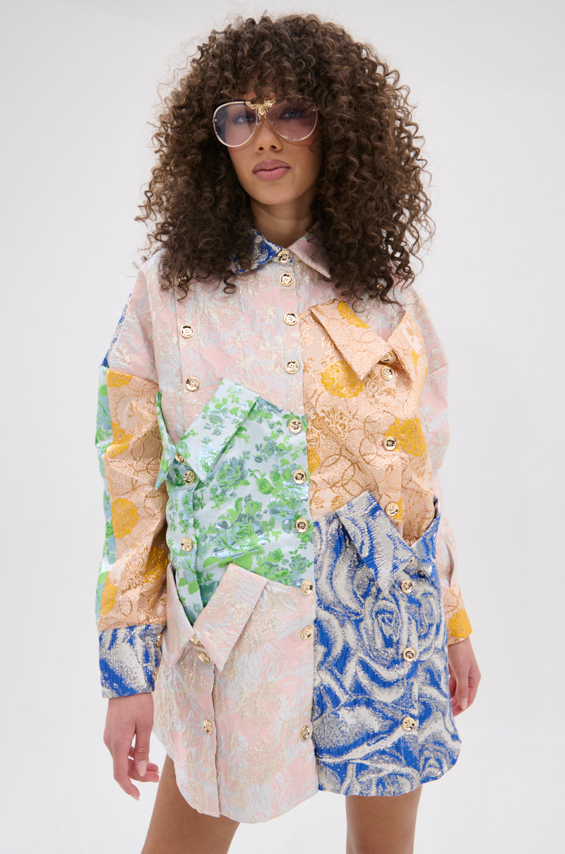 A woman with curly hair and sunglasses wears the MY GIRLFRIENDS SHIRT BROCADE MINI DRESS, featuring pastel floral patchwork, metallic buttons, and oversized pockets, standing against a plain light background.