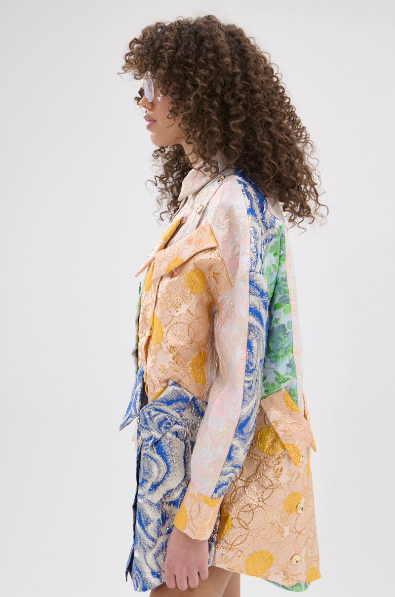 A person with curly hair and sunglasses models the MY GIRLFRIENDS SHIRT BROCADE MINI DRESS, featuring a colorful patchwork design with blue, green, and orange sections, standing sideways against a plain light background.