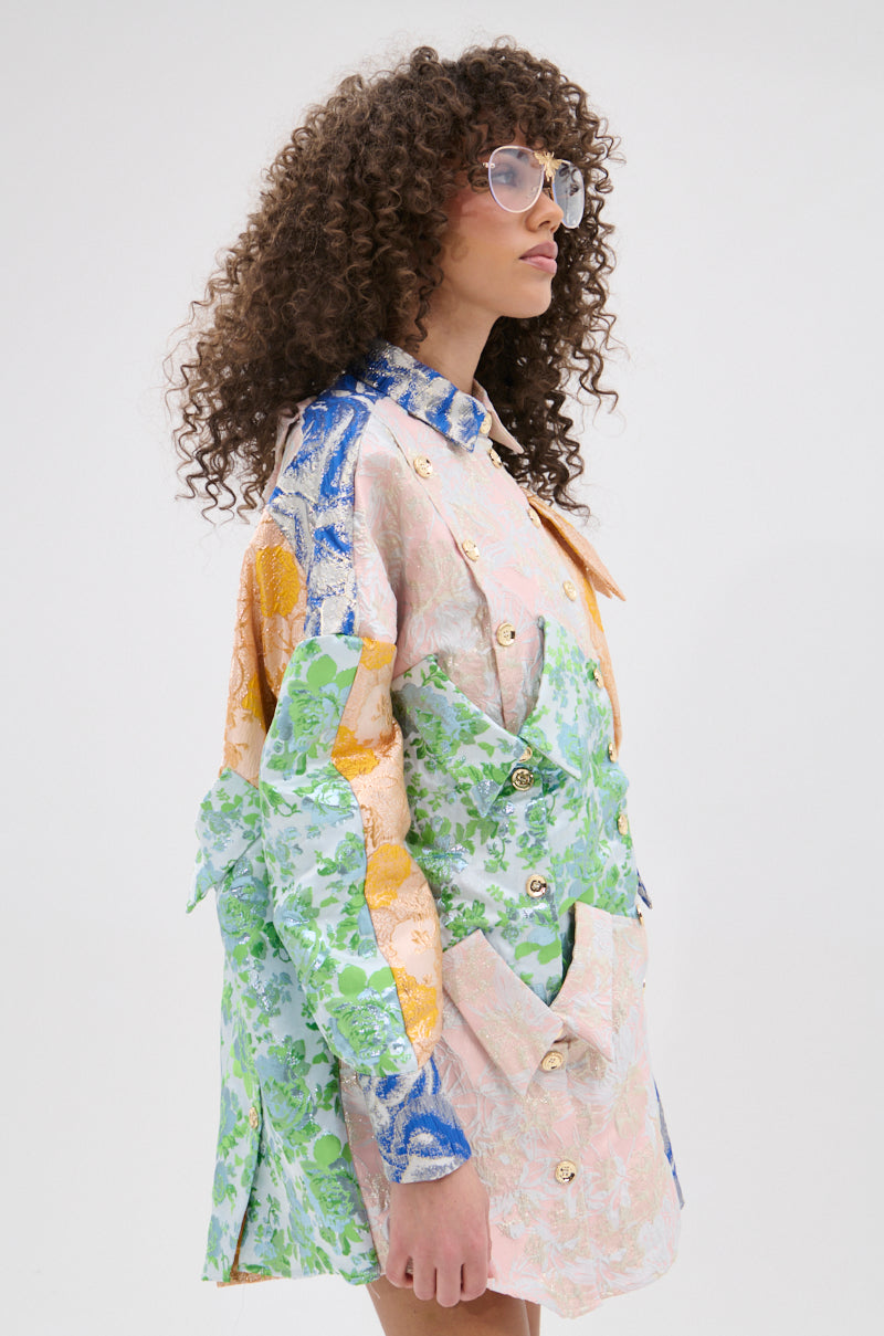 A person with curly hair and large sunglasses stands against a plain white background wearing the MY GIRLFRIENDS SHIRT BROCADE MINI DRESS, which features metallic buttons and pastel green, blue, yellow, and pink floral designs.