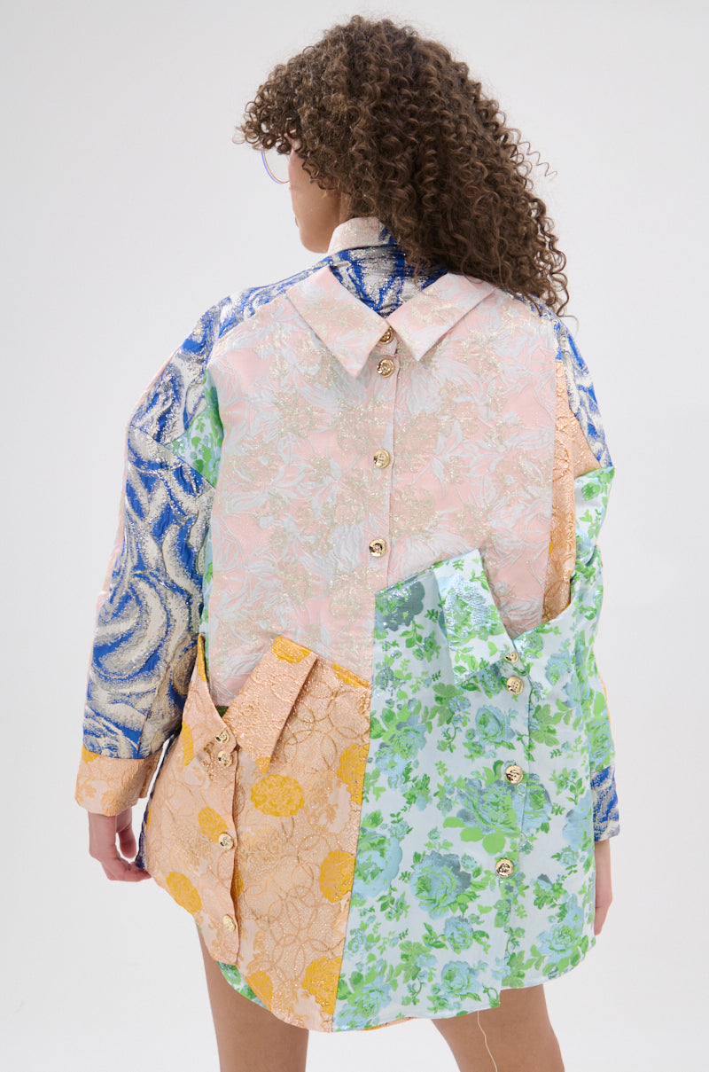 A woman with curly hair faces away, wearing the MY GIRLFRIENDS SHIRT BROCADE MINI DRESS—a patchwork shirt-dress in blue, pink, yellow, and green fabrics with collar accents and metallic button details.