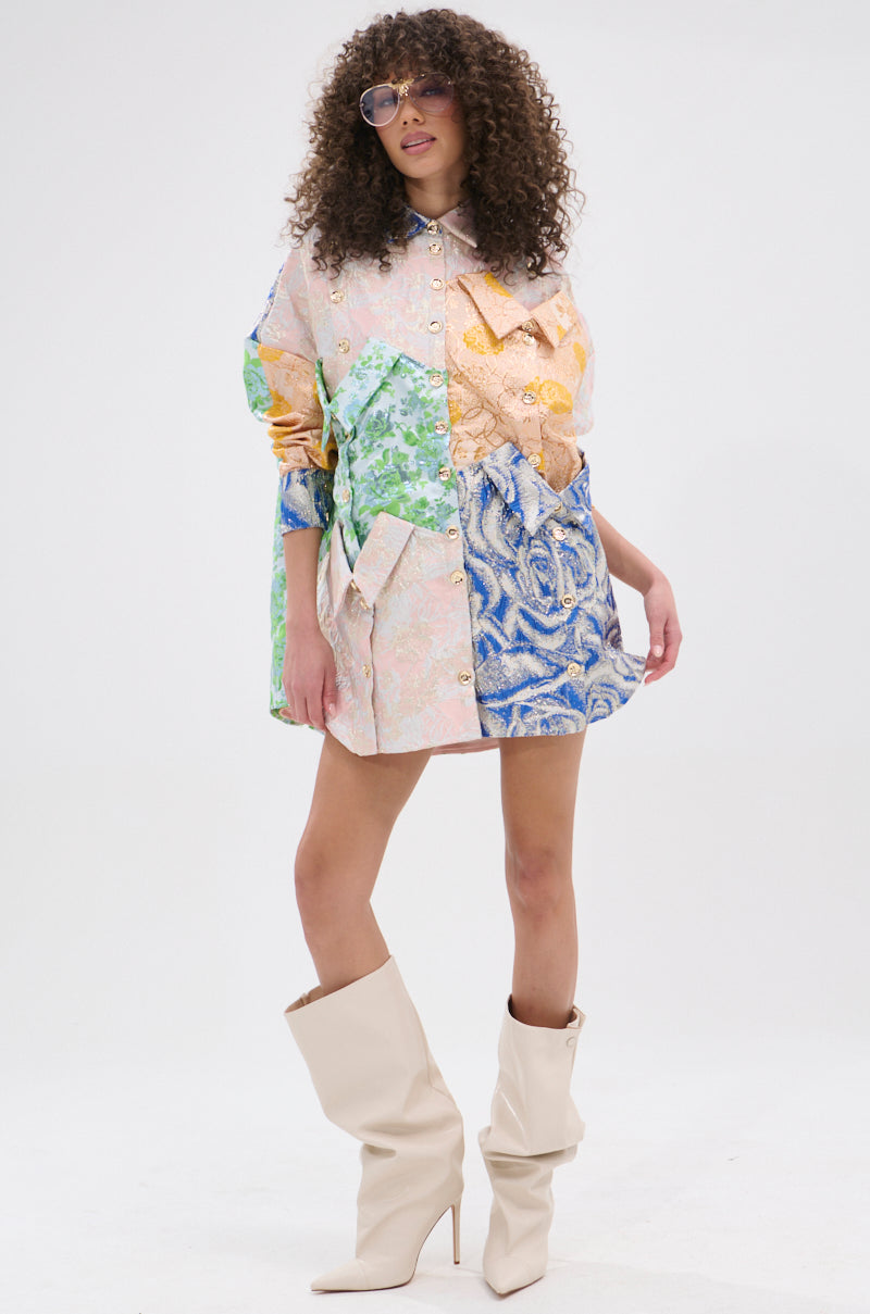 A woman with curly hair wears sunglasses and the MY GIRLFRIENDS SHIRT BROCADE MINI DRESS, featuring a colorful patchwork design, large pockets, and metallic buttons, paired with slouchy cream knee-high boots against a plain white background.