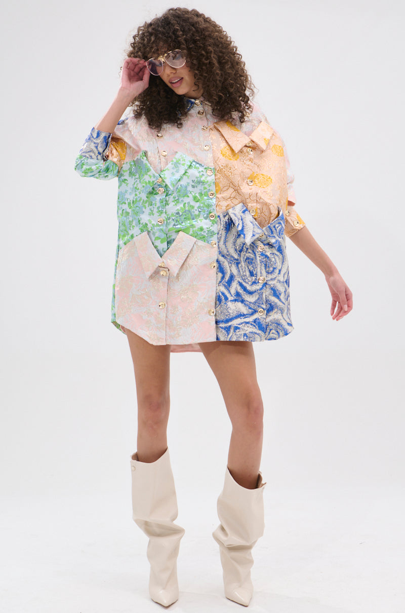 A woman with curly hair in oversized sunglasses models the MY GIRLFRIENDS SHIRT BROCADE MINI DRESS, a patchwork shirt-dress with metallic buttons and pastel floral patterns, paired with knee-high off-white boots against a white background.