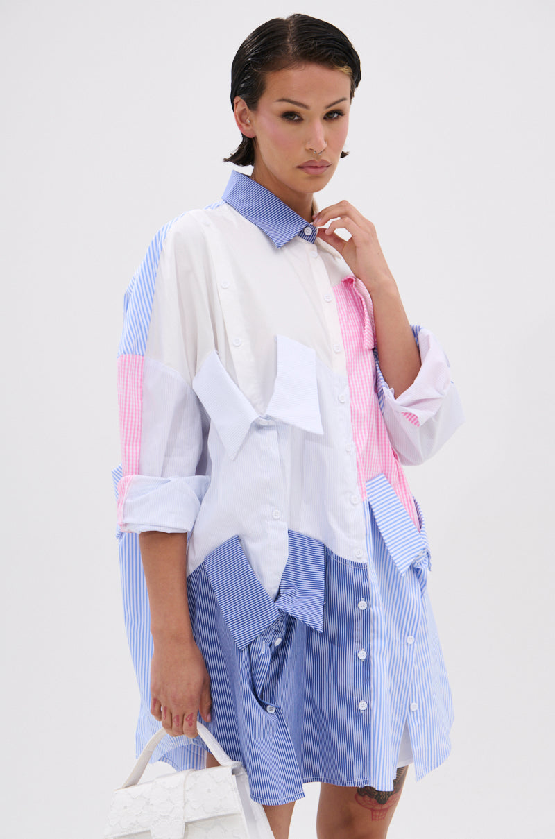 A woman with slicked-back hair and a neutral expression stands against a plain white background, wearing the MY BOYFRIENDS SHIRT POPLIN MINI DRESS in oversized white, blue, and pink stripes and holding a textured white handbag.