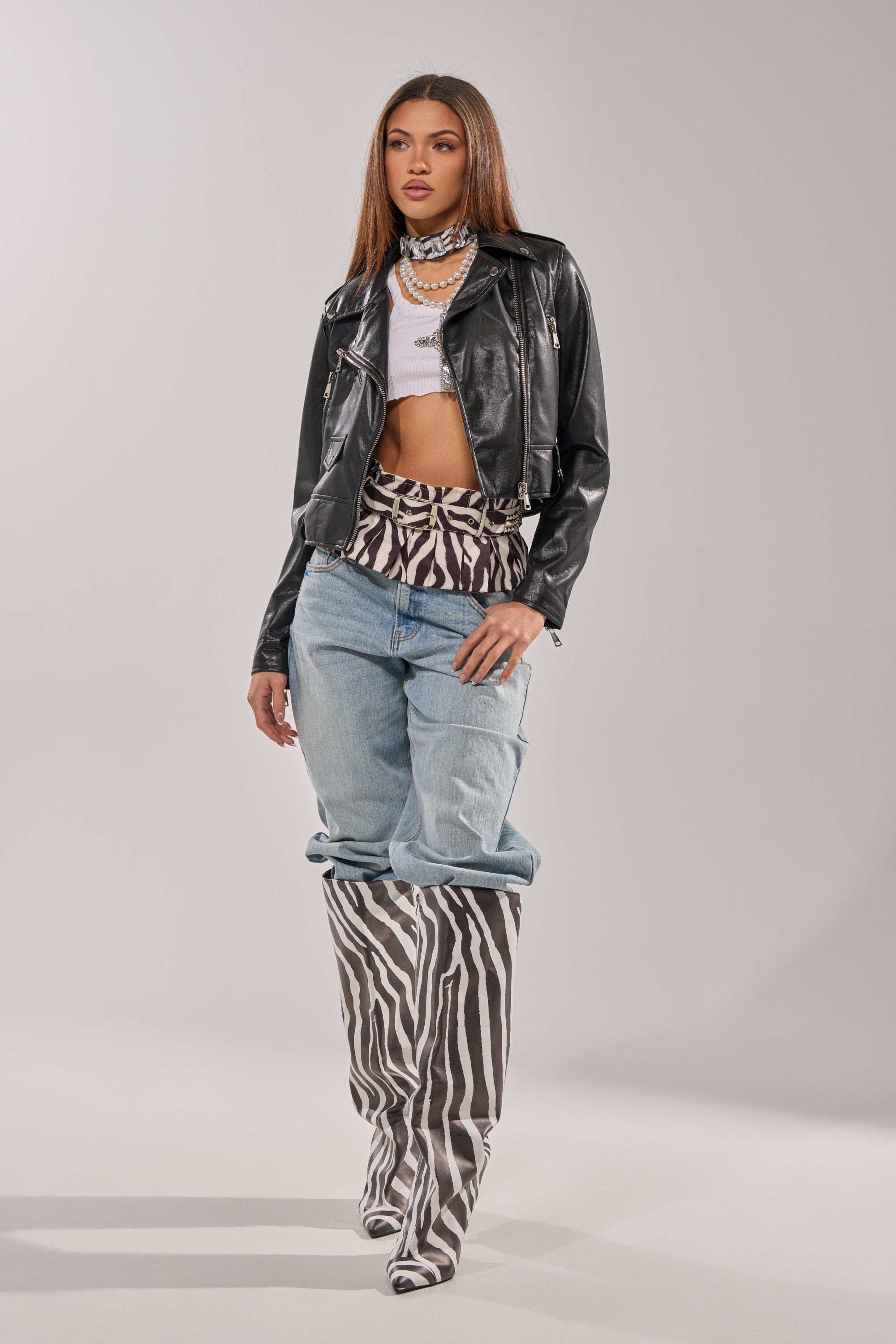 A woman poses against a plain light background wearing a black leather jacket, white cropped top, patterned scarf, layered necklaces, and the ZEBRA PEPLUM BELT SKIRT.