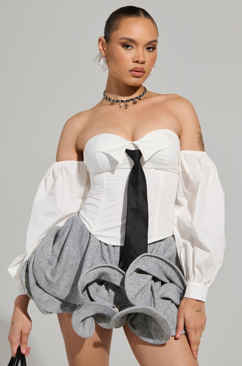 Wearing the LETS DO IT BLOUSE, a person poses confidently in an off-the-shoulder design with a tie detail, paired perfectly with puffy gray shorts. Their hair is styled back, accentuated by a choker and earrings. A black bag completes the look against a simple background.