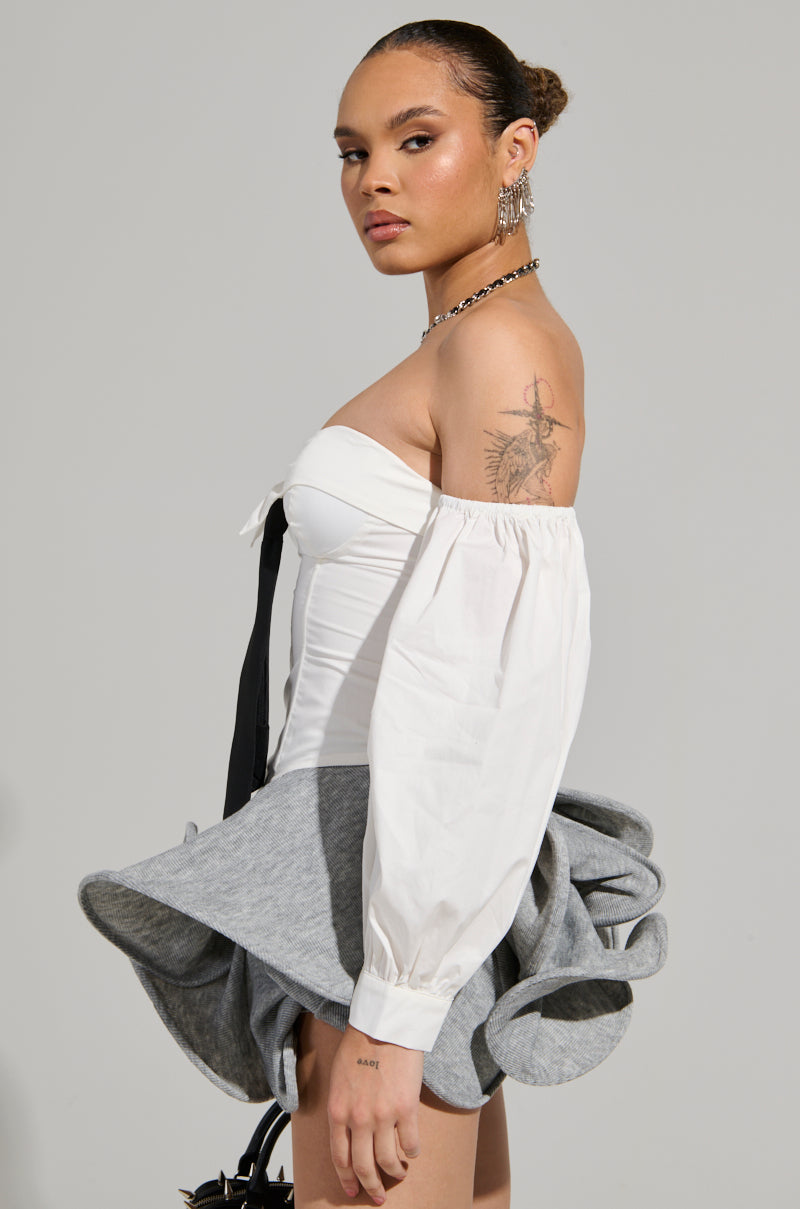 A person stands confidently against a neutral background, wearing the chic LETS DO IT BLOUSE and a unique gray skirt. They showcase a tattoo on their arm, along with several earrings and a necklace. Their hair is styled elegantly in a bun.