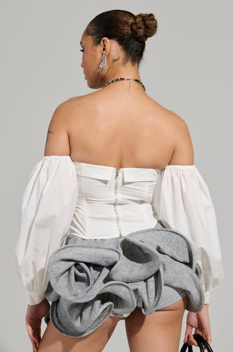 A person wearing the LETS DO IT BLOUSE, which is a white off-the-shoulder design featuring puff sleeves, pairs it with an elaborate gray skirt and stands with their back to the camera. They accessorize with chain earrings, a necklace, and hold a small black bag. Their hair is elegantly styled in a bun.