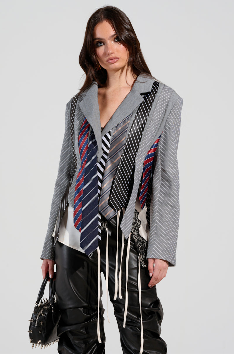 A woman showcases the STRICTLY BUSINESS TIE DETAIL BLAZER, crafted from various neckties over a grey pinstripe base, complemented by black leather pants. She clutches a studded black handbag. Her hair flows freely as she stands against a plain background, exuding effortless style.