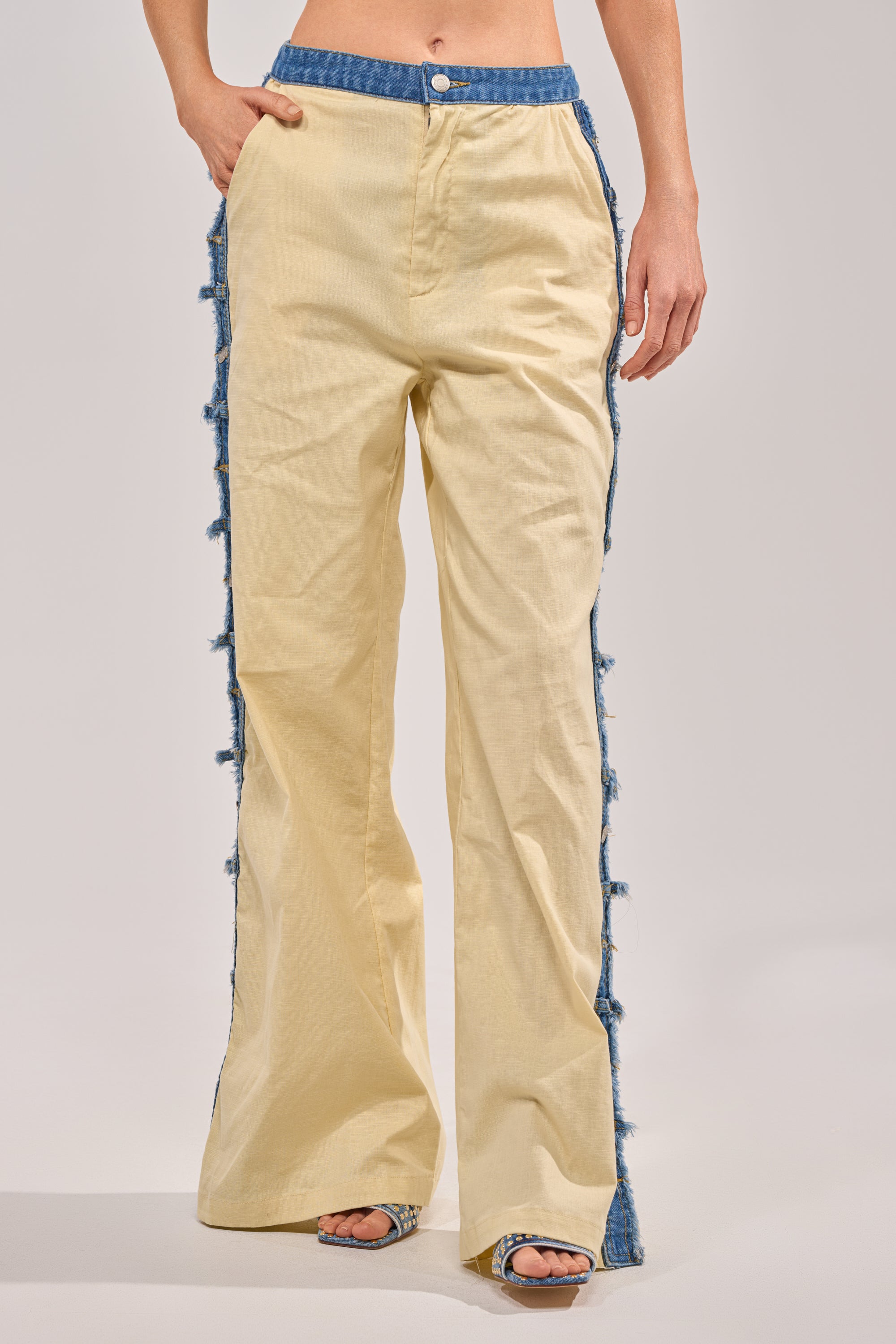 Someone wearing the DO YOU LOVE ME TROUSER—high-waisted, wide-leg beige linen pants with denim trim, frayed side edges, and a button-zip closure with denim waistband accents—paired with open-toe heels.