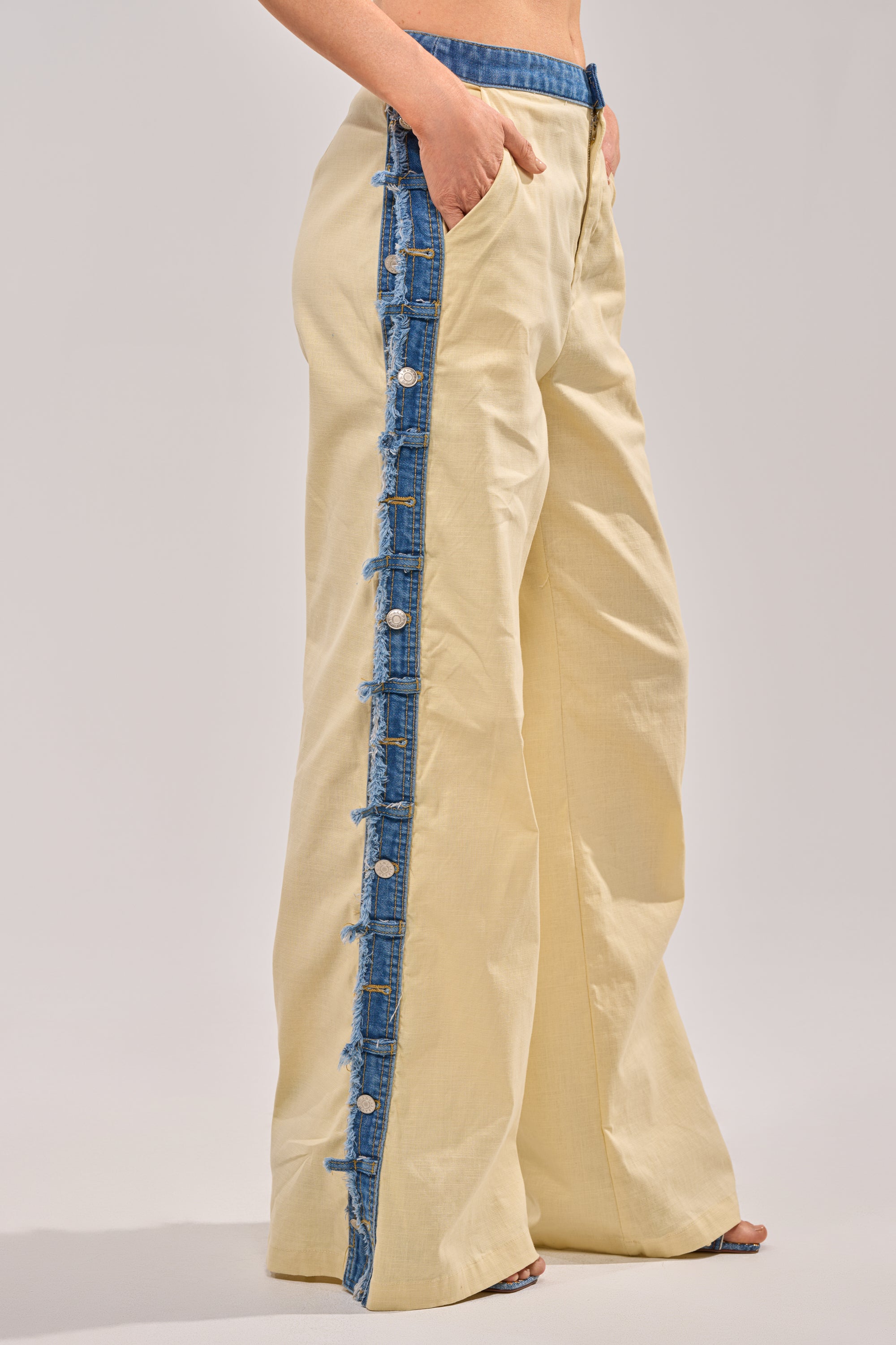 A person wears the DO YOU LOVE ME TROUSER: wide-leg, light yellow linen pants with blue denim trim, prominent buttons, raw edges, and pockets. The model stands against a plain background with one hand in a pocket.