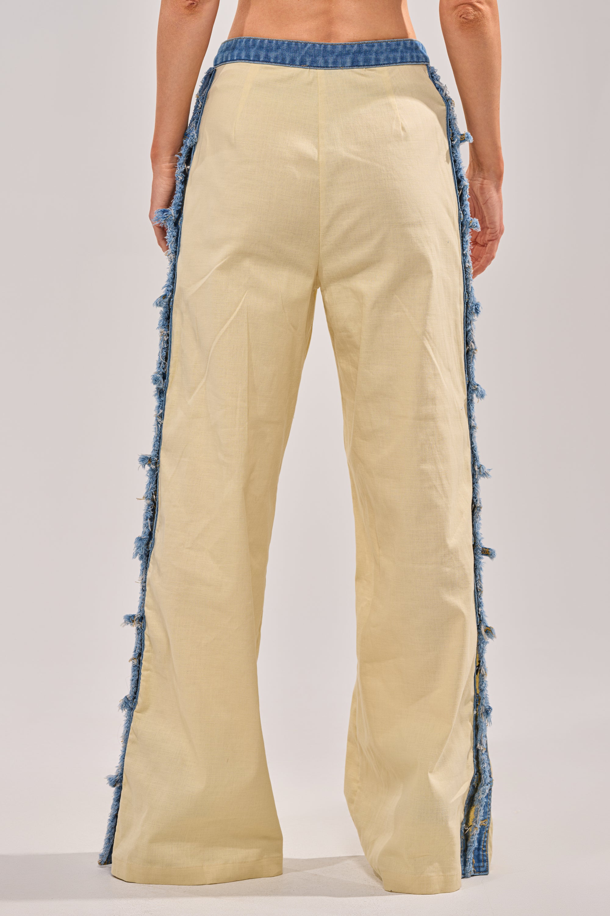 A person shown from the back wears the DO YOU LOVE ME TROUSER—a pair of high-waisted, wide-leg cream linen pants featuring blue denim trim along the side seams—posed against a plain background.