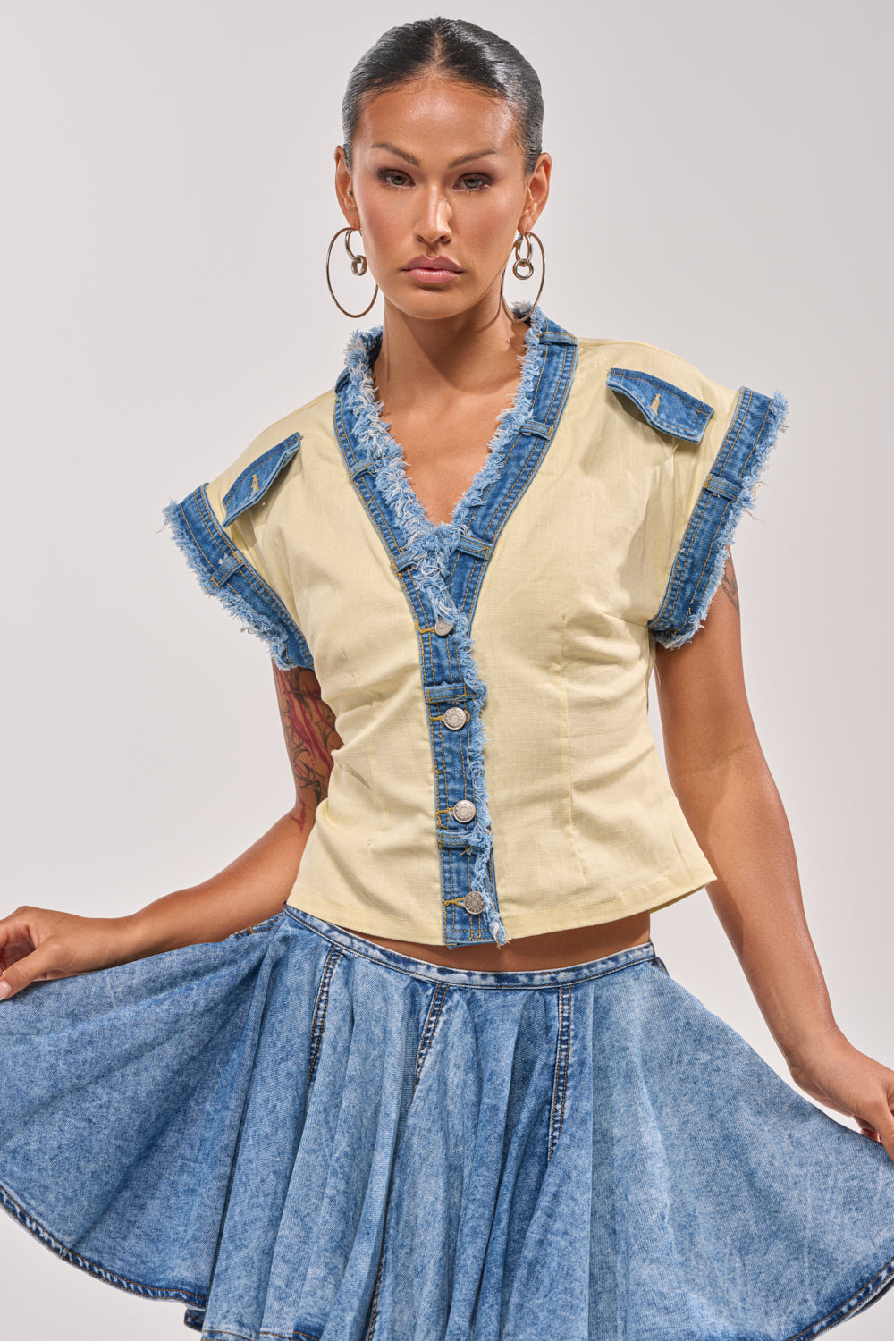 A woman wears the DO YOU LOVE ME BUTTON DOWN, a yellow top with denim trim and frayed edges, paired with a blue denim skirt. She accessorizes with hoop earrings, pulls her hair back, and holds the edge of her skirt. 0