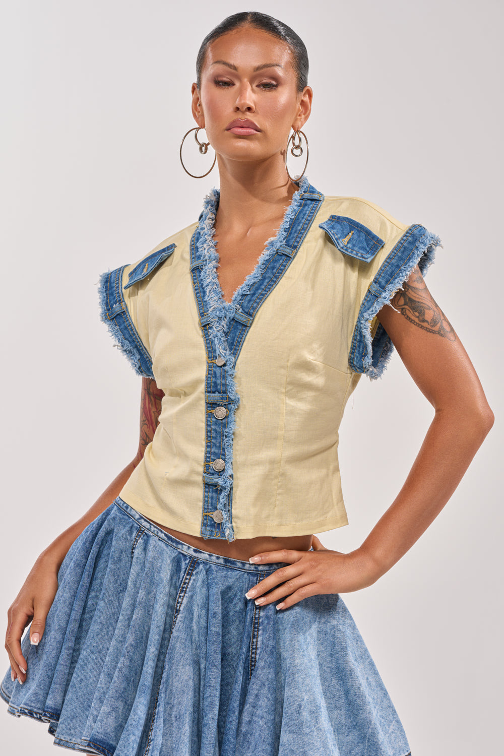 A woman confidently poses against a plain background wearing the DO YOU LOVE ME BUTTON DOWN, a v-neck yellow top with blue buttons and denim trim, paired with a flared blue denim skirt. She accessorizes with hoop earrings and showcases her tattoos. 1