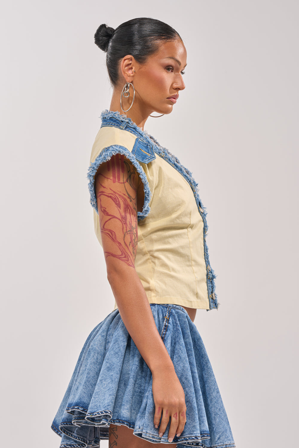 Woman with dark hair in a bun, large hoop earrings, and a blue denim skirt stands in profile against a plain backdrop. She pairs her look with the DO YOU LOVE ME BUTTON DOWN, a beige sleeveless top with frayed denim details. 2