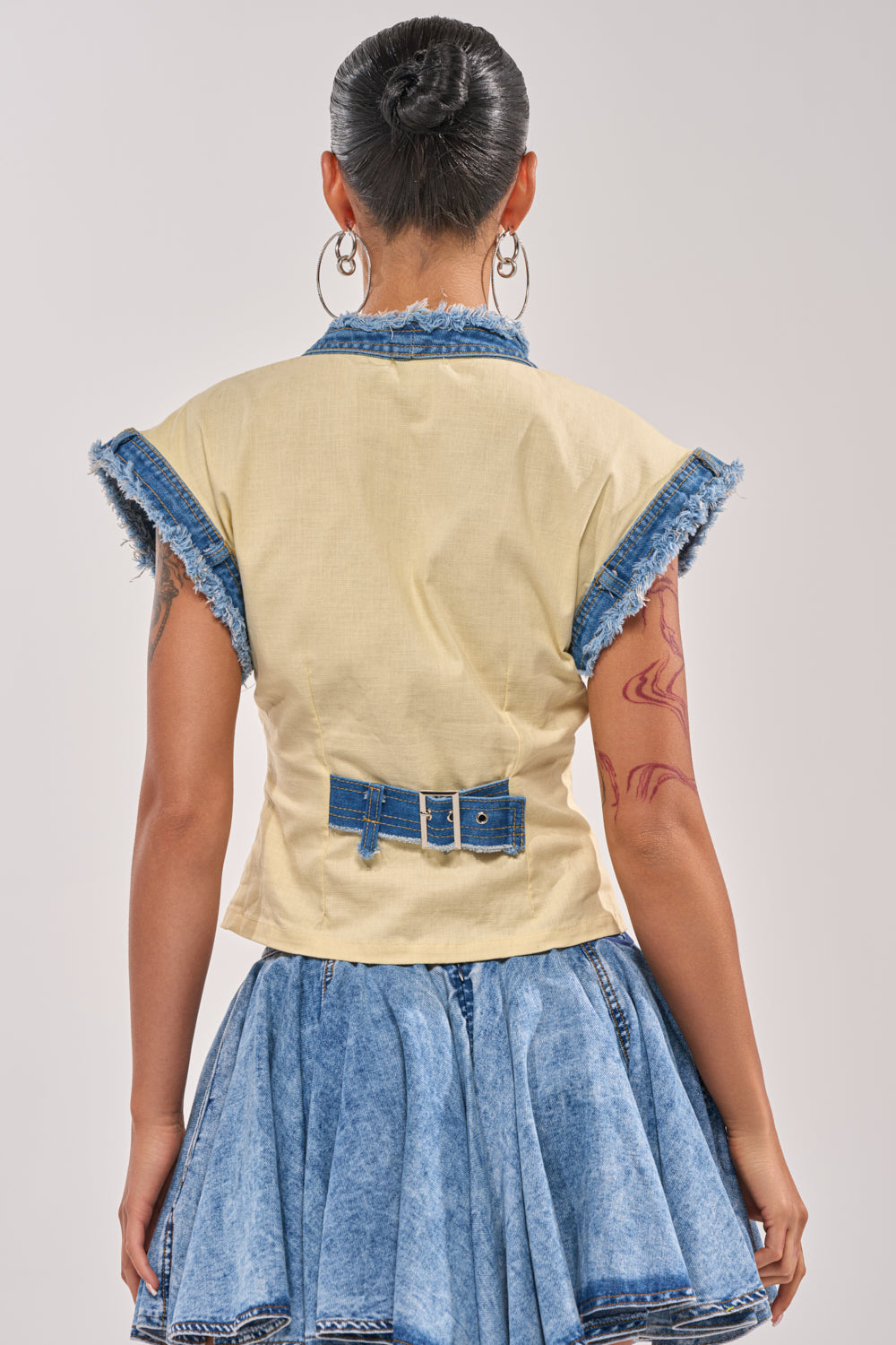 A person with dark hair in a bun is seen from behind, wearing the DO YOU LOVE ME BUTTON DOWN—a short-sleeved yellow top with denim trim, button details, and a blue denim belt—paired with a flared blue denim skirt. 3