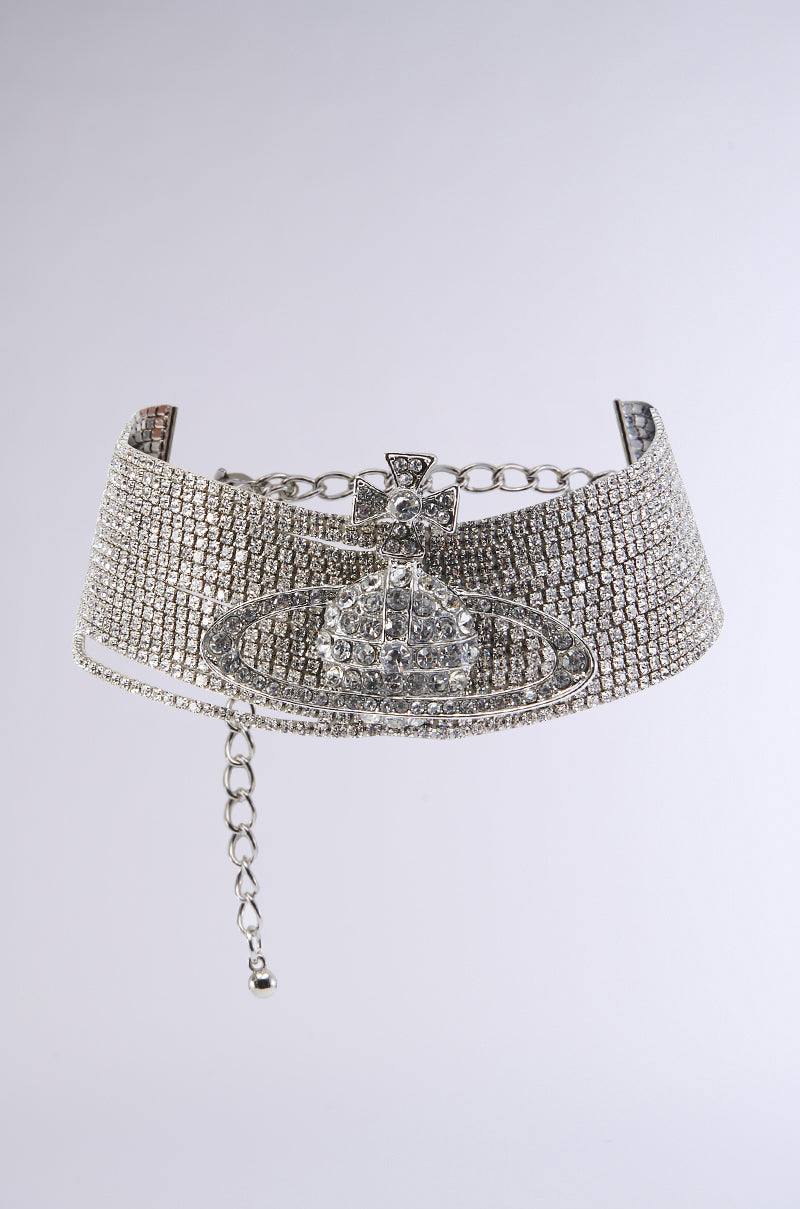 A close-up image of the TITI CHOKER NECKLACE showcases its sparkling rhinestone chain links. The centerpiece is a prominent, intricate emblem featuring a cross enclosed in an oval. This shiny, silver choker includes an adjustable lobster clasp closure and a dangling end for fastening. The background is plain and light gray.
 0