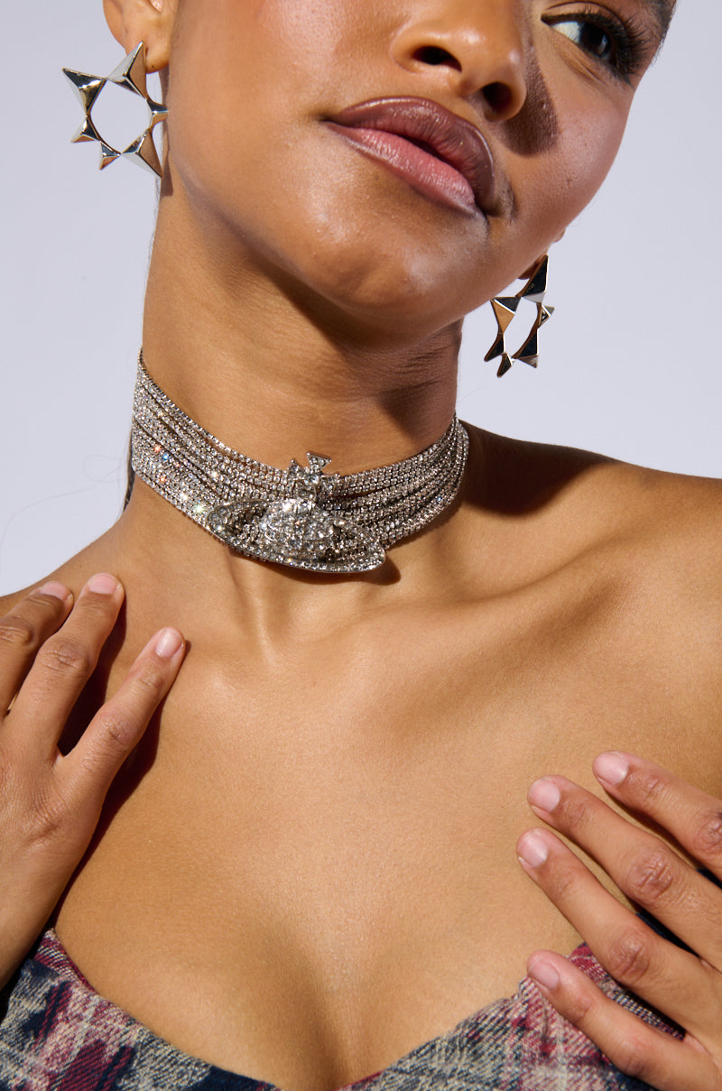 A close-up of a person wearing geometric hoop earrings and a sparkling TITI CHOKER NECKLACE with rhinestone chain links. The person is gently touching their neck with one hand, highlighting the adjustable lobster clasp closure, while part of their upper outfit showcases a textured fabric pattern. 1