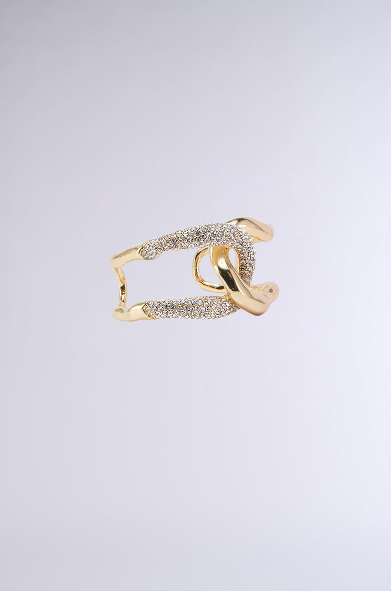 The INTERTWINED BRACELET CUFF features a gold and silver design with a twisted knot motif. The gold band intertwines with a silver band adorned with pave rhinestone details, creating an elegant, intertwined look against a plain, light grey background.