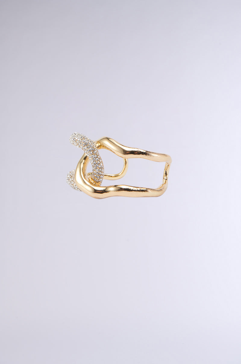 The INTERTWINED BRACELET CUFF is a faux gold metallic piece featuring an abstract design with intertwining loops, one of which is adorned with small, sparkling white gemstones. The bracelet boasts pave rhinestone detailing and is displayed against a plain light gray background.