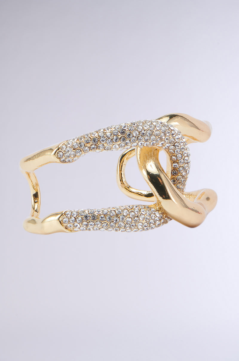 A close-up image of the INTERTWINED BRACELET CUFF in opulent faux gold metallic, showcasing its unique intertwined design. The bracelet is adorned with pave rhinestone detailing and numerous sparkling white crystals, resulting in a luxurious and elegant appearance. The background features a soft gradient of light grey and white.