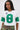 A woman wears the WINNERS ONLY RHINESTONE SHORT SLEEVE TEE, styled as a varsity graphic white crop top football jersey with a large green number 8, green sleeve stripes, camouflage pants, long straight hair, and hoop earrings against a plain background.
