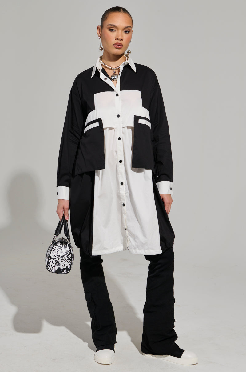 A person stands against a plain background wearing the NOT FEELIN IT PARACHUTE MIDI DRESS, featuring a long button-up style with a black-and-white color-block design and large pockets. They carry a small patterned bag and accessorize with layered necklaces, completing the look with black pants and white shoes. 2