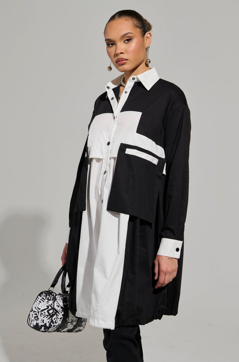 A person wearing the NOT FEELIN IT PARACHUTE MIDI DRESS, characterized by its black-and-white geometric design, poses confidently. They hold a small patterned handbag and wear earrings. The plain gray background further enhances this striking fashion ensemble. 3