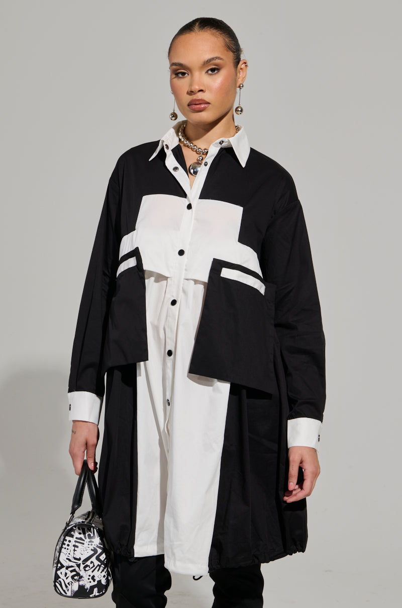 The person is wearing the NOT FEELIN IT PARACHUTE MIDI DRESS, which features a stylish black and white button-down design with layered fabric and pockets, resembling a parachute midi dress. They have accessorized with jewelry and are holding a small patterned handbag, posing against a plain background. 1