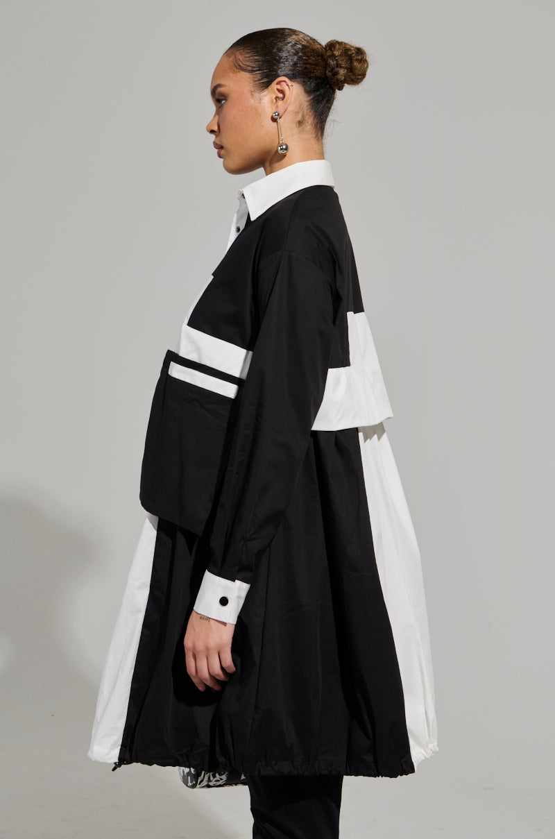 A person poses in profile, dressed in the NOT FEELIN IT PARACHUTE MIDI DRESS, featuring a chic black and white color-block design with prominent large pockets. Their hair is elegantly tied back in a bun, and the plain light gray background accentuates the dress’s windbreaker-inspired elements. 4