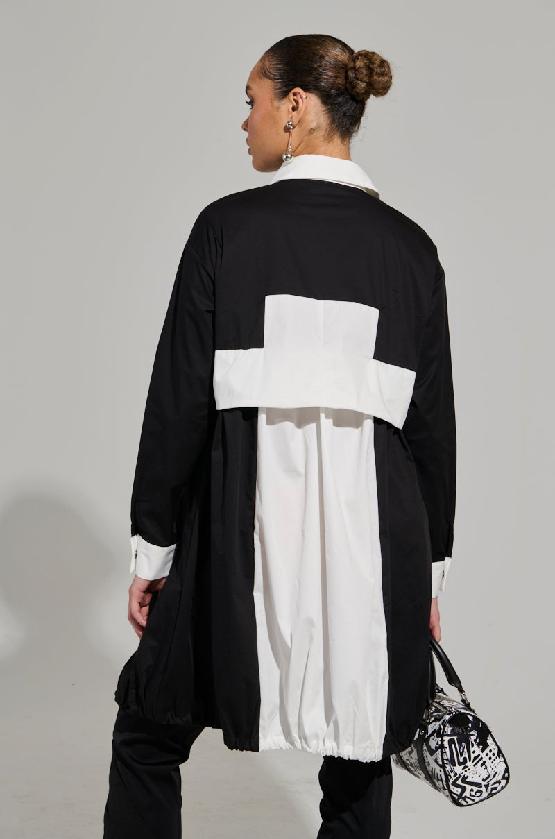 A person with hair in a bun wears the NOT FEELIN IT PARACHUTE MIDI DRESS, featuring a black and white long coat design with a white shirt collar and cuffs, evoking a windbreaker-inspired style. They hold a patterned handbag while standing slightly away from the camera against a plain light background. 5