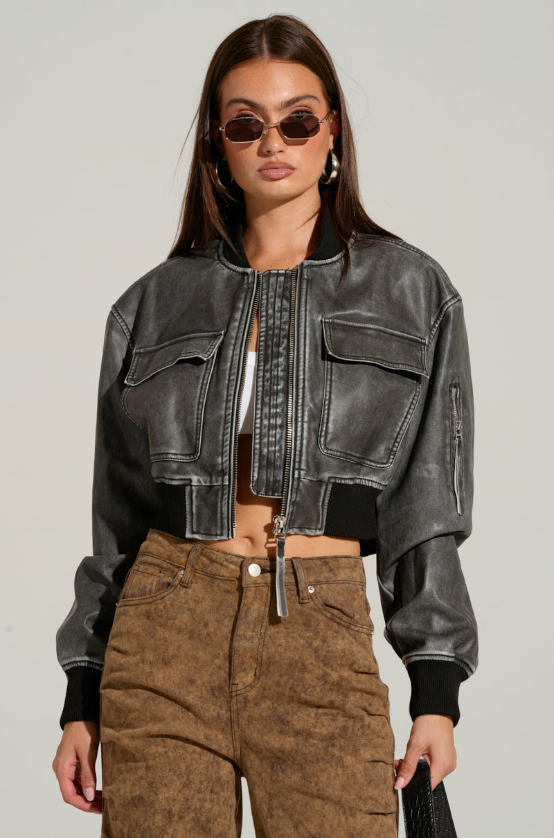 A woman with long brown hair stands against a plain background wearing a cropped Lunar Mineral Wash Bomber with a metallic front zipper closure, light brown high-waisted pants, and sunglasses. She also wears large hoop earrings and holds a black bag.