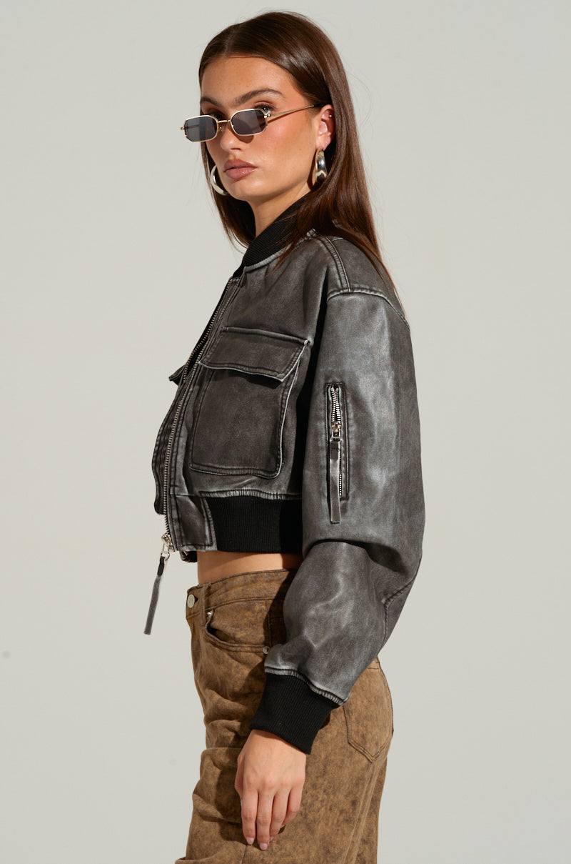 A woman with long brown hair and sunglasses is wearing the LUNAR MINERAL WASH BOMBER, a cropped gray faux leather jacket with multiple pockets and a metallic front zipper closure. She pairs it with brown pants, posing sideways and looking at the camera over her shoulder.