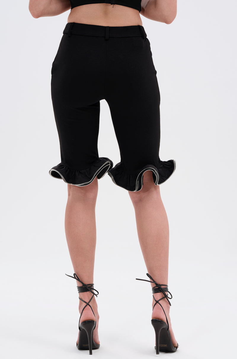 A person faces away, wearing the DAINTY DETAILS RUFFLE HEM TAILORED SHORT in black with high-heeled ankle strap sandals against a plain white background.