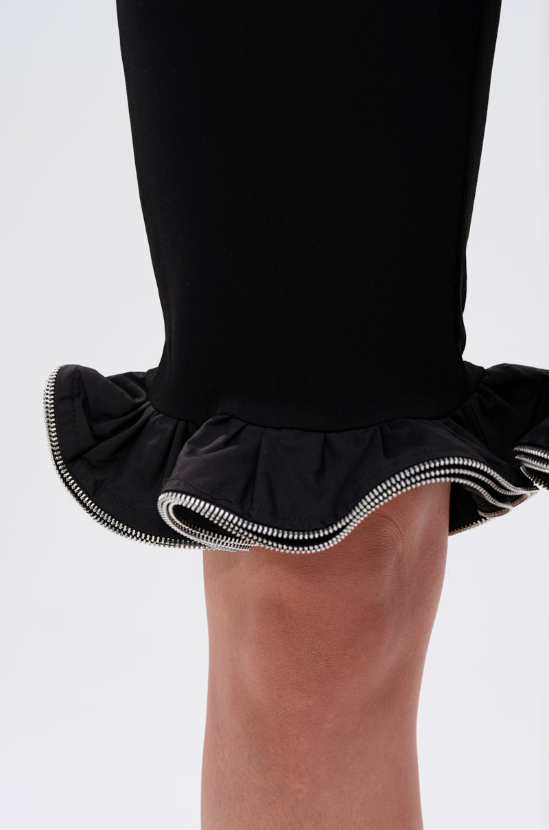 A close-up shows a leg wearing the DAINTY DETAILS RUFFLE HEM TAILORED SHORT in black, featuring a high rise fit, ruffle hem, and decorative white zipper trim against a plain light background.