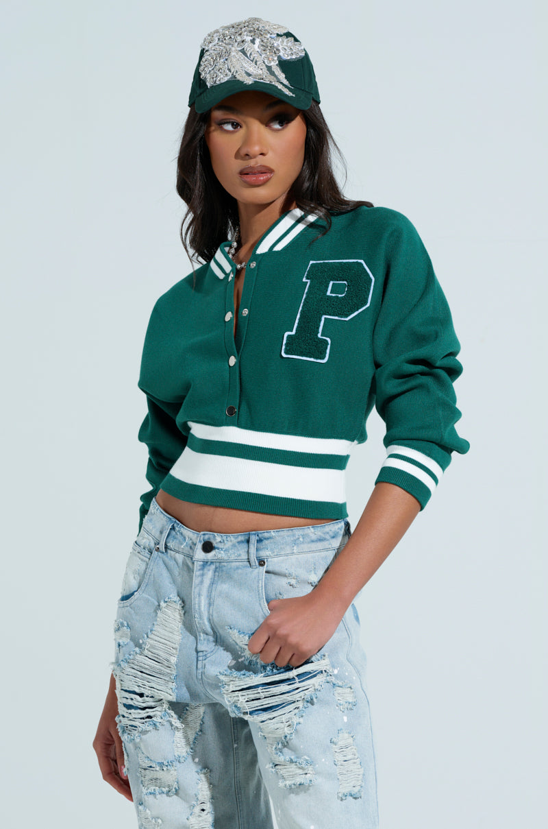 Wearing a VARSITY TEAM BUTTON DOWN SWEATER with a large "P" on the front and a sequined cap, someone pairs the sporty top with distressed light blue jeans, posing confidently against a plain background. 1