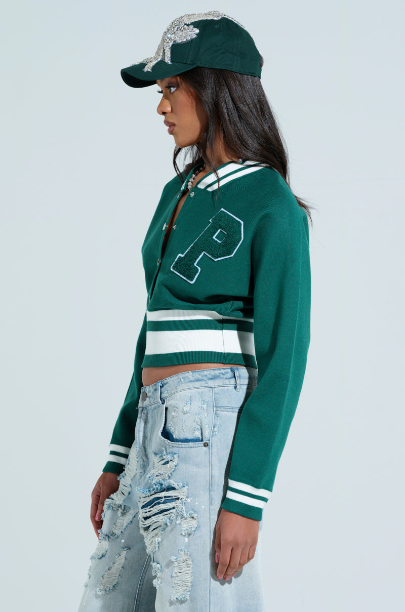 The woman showcases a cropped look with her green VARSITY TEAM BUTTON DOWN SWEATER featuring a letter "P," paired with distressed light blue jeans and a dark green, sequin-studded cap. Against a plain backdrop, she exudes understated style, facing sideways. 2