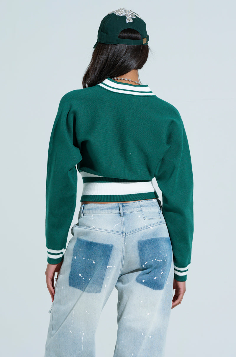 A person sports a green VARSITY TEAM BUTTON DOWN SWEATER and matching cap with a white design on the back. They have long hair, light blue jeans with dark patches and white paint splatters, capturing a sporty-chic vibe from behind. 3