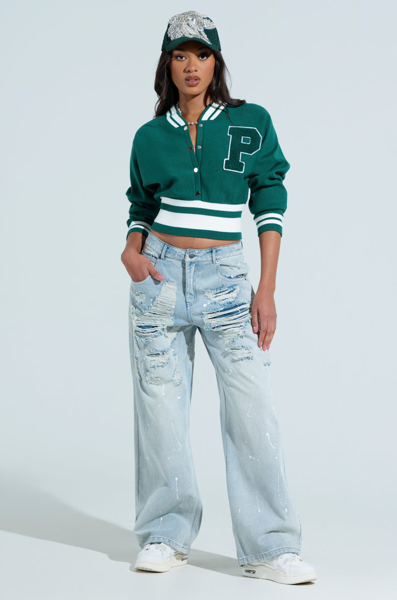 Dressed in a green varsity team button-down sweater with a "P" emblem, paired with a matching cap and light blue distressed jeans, they exude sporty-chic style. Their white sneakers perfectly complete the ensemble against a plain light gray background. 4