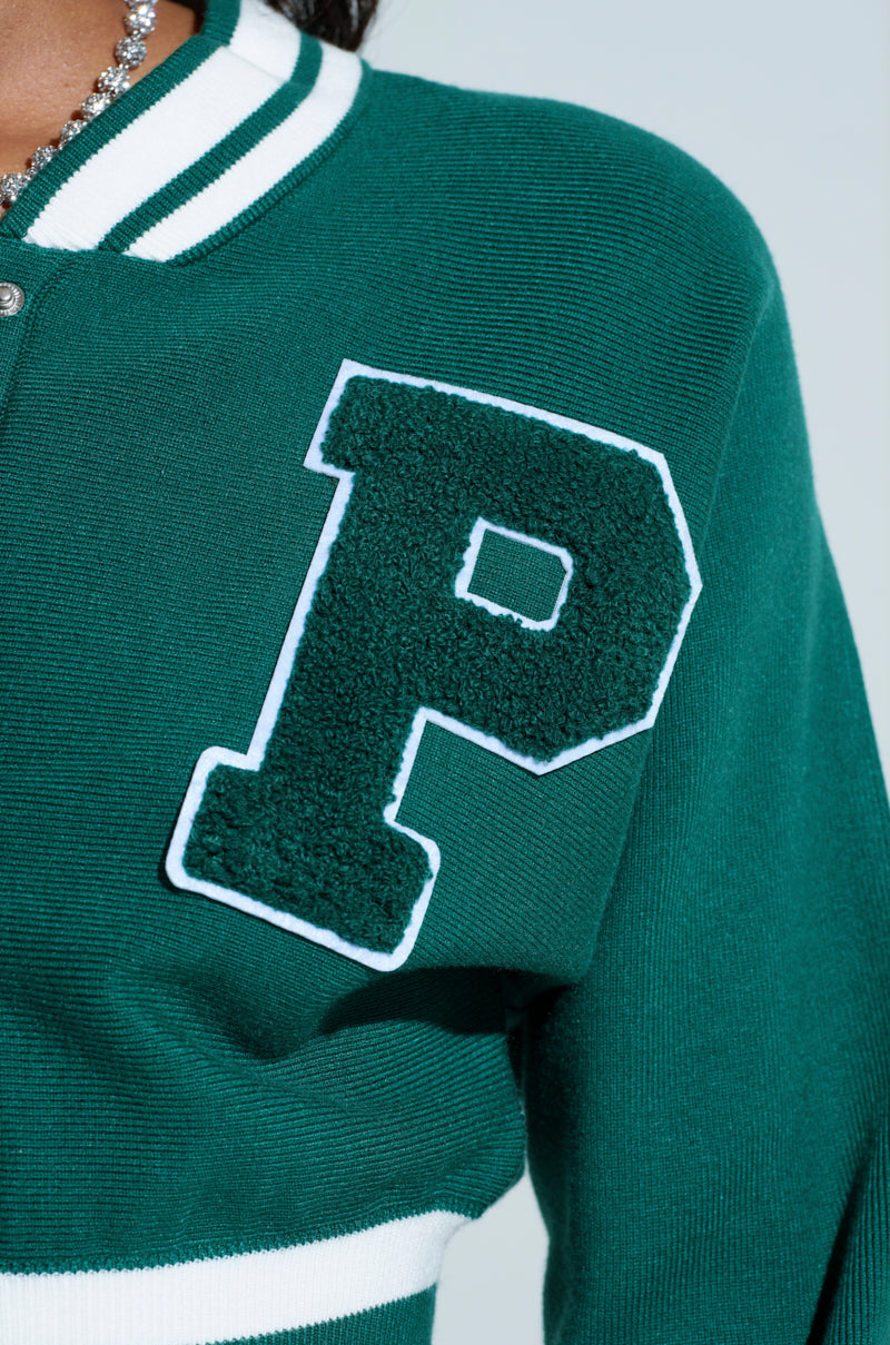 A person sports the Varsity Team Button Down Sweater, a green varsity-style jacket with a large textured "P" patch on the chest, exuding sporty-chic vibes. White trim along the collar enhances its stylish flair. 5