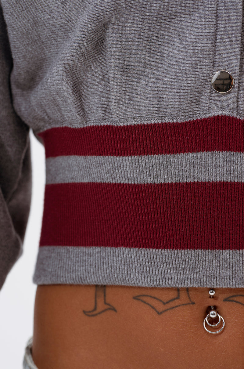 Close-up of a person wearing the VARSITY TEAM BUTTON DOWN SWEATER IN HEATHER GREY, featuring red and gray stripes at the cropped hem. The lower torso shows a geometric tattoo and two silver belly button piercings. 9