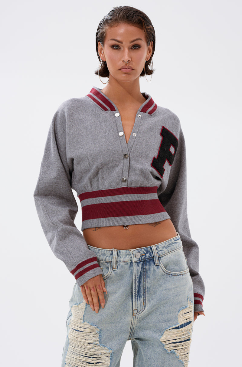 A person wears the Varsity Team Button Down Sweater in heather grey, featuring red stripes and a letter "P," paired with light blue distressed jeans. They have slicked-back hair and a neutral expression. 1