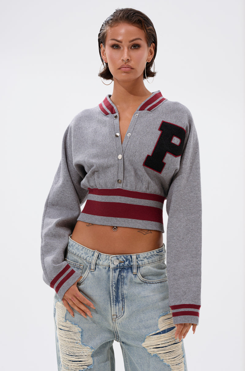 A woman models the Varsity Team Button Down Sweater in Heather Grey, featuring red stripes and a large letterman patch, styled with light blue distressed jeans against a plain white background. 2