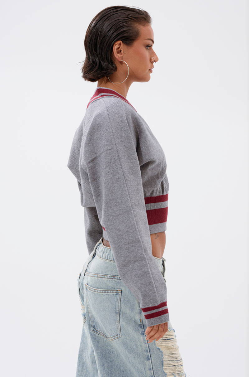 A woman with short dark hair and large hoop earrings wears the VARSITY TEAM BUTTON DOWN SWEATER IN HEATHER GREY, styled with distressed light blue jeans, standing in profile against a plain white background. 3