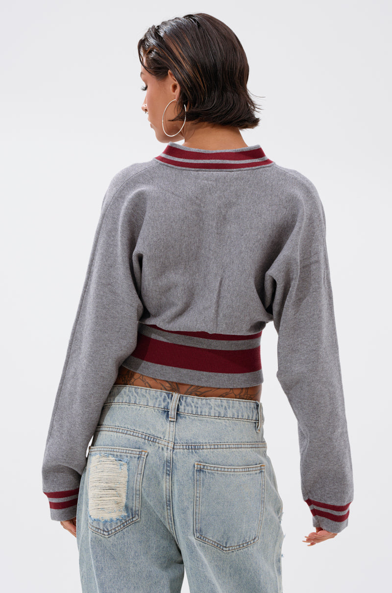 A person with short dark hair wears large hoop earrings and the VARSITY TEAM BUTTON DOWN SWEATER IN HEATHER GREY, styled with light blue distressed jeans, standing facing away from the camera against a plain light background. 5